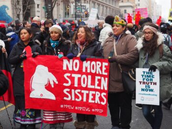 A group of indigenous women confidently walk down a city street while holding a red banner that reads: "no more stolen sisters." They are at a crowded protest. They are wearing coats, hats and gloves.